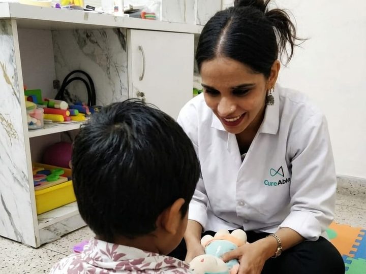 A smiling therapist sitting on a play mat, offering a child a soft toy.