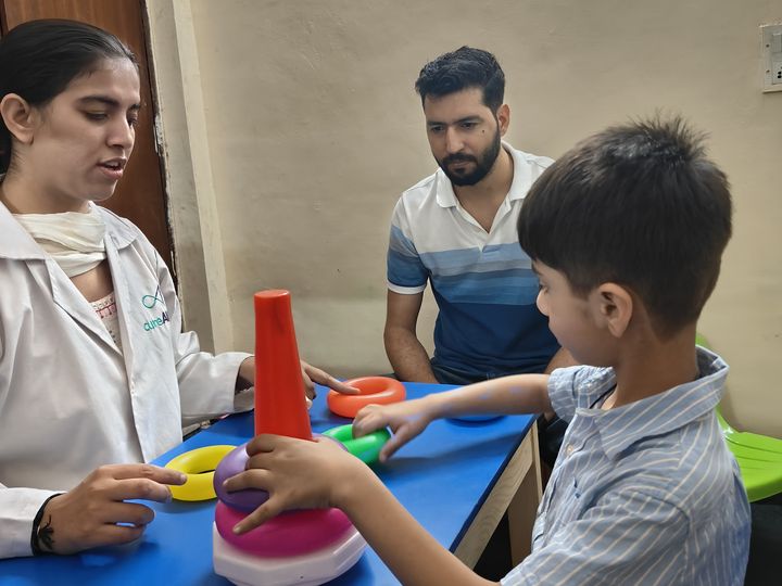 A child stacking rings at a table with a therapist while an adult watches.
