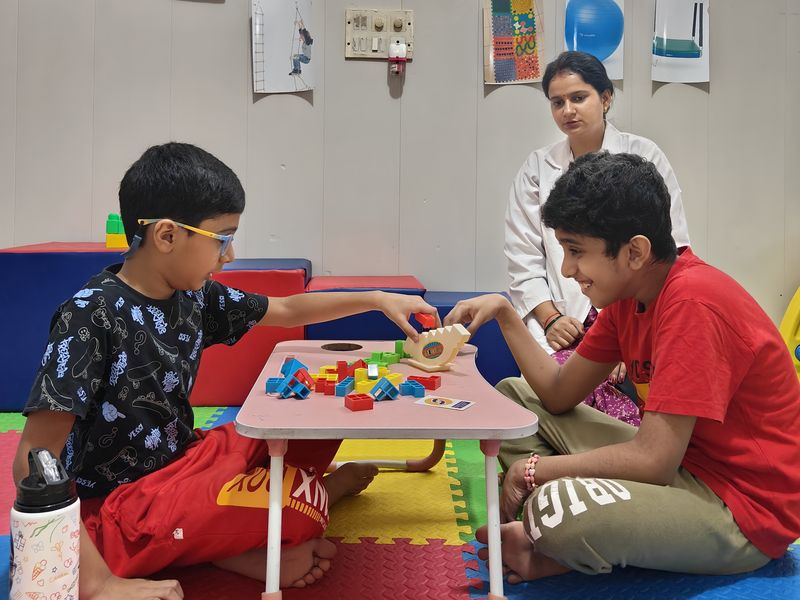 Two children playing with building blocks together while a therapist watches.