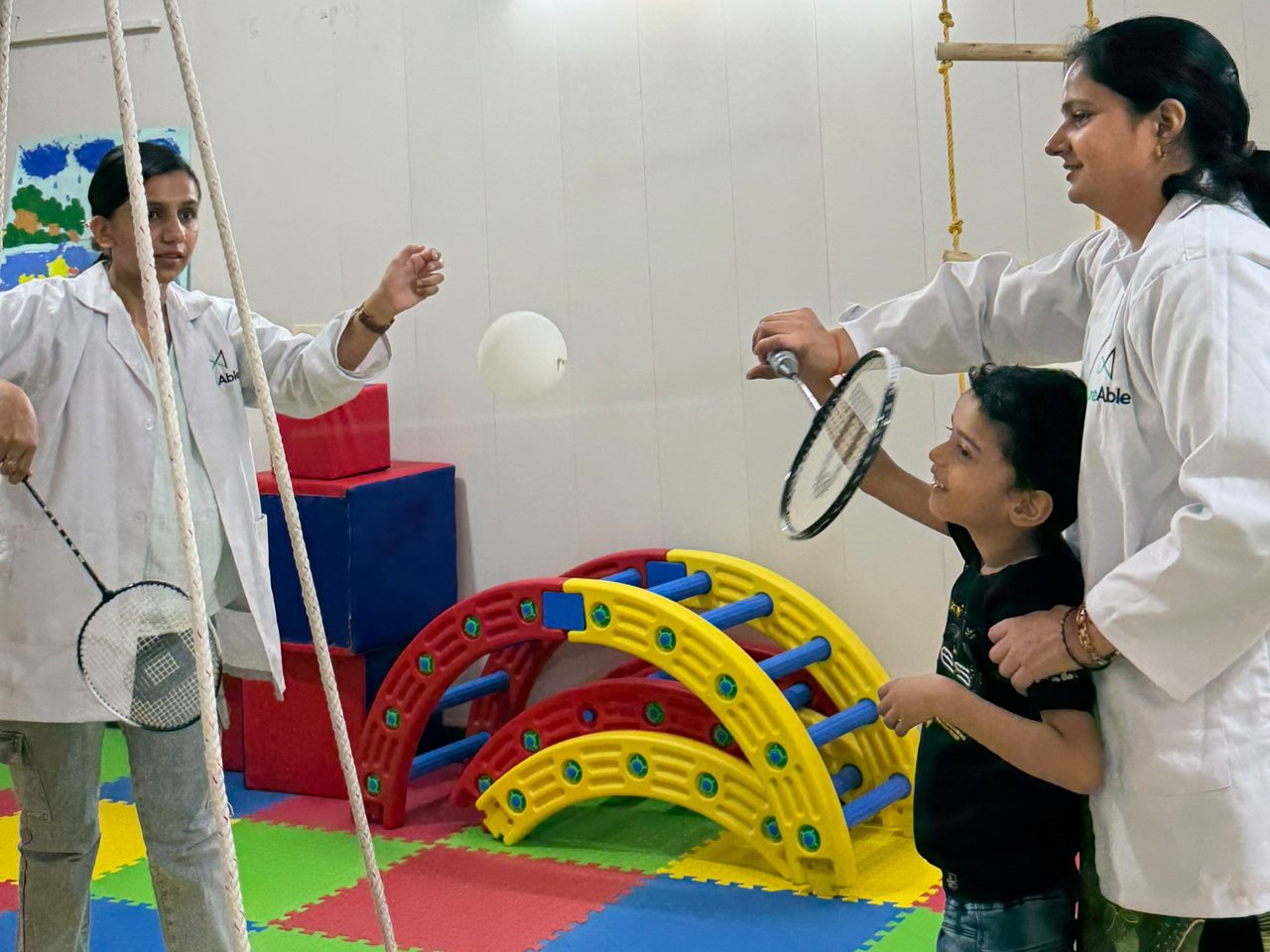 Two CureAble team members playing a racket-and-balloon game with a child in a therapy room.