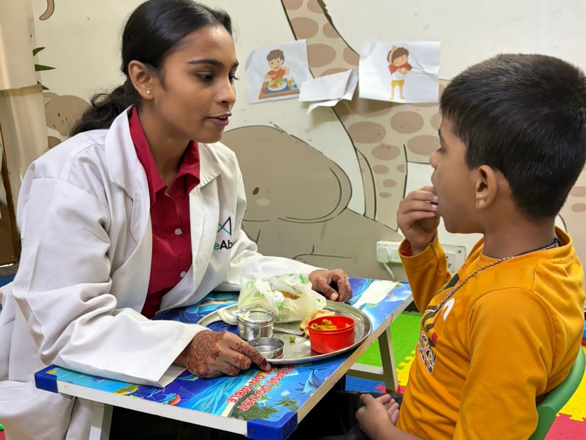 A CureAble therapist sitting with a child at a small table during a mealtime activity at the Dwarka Centre.