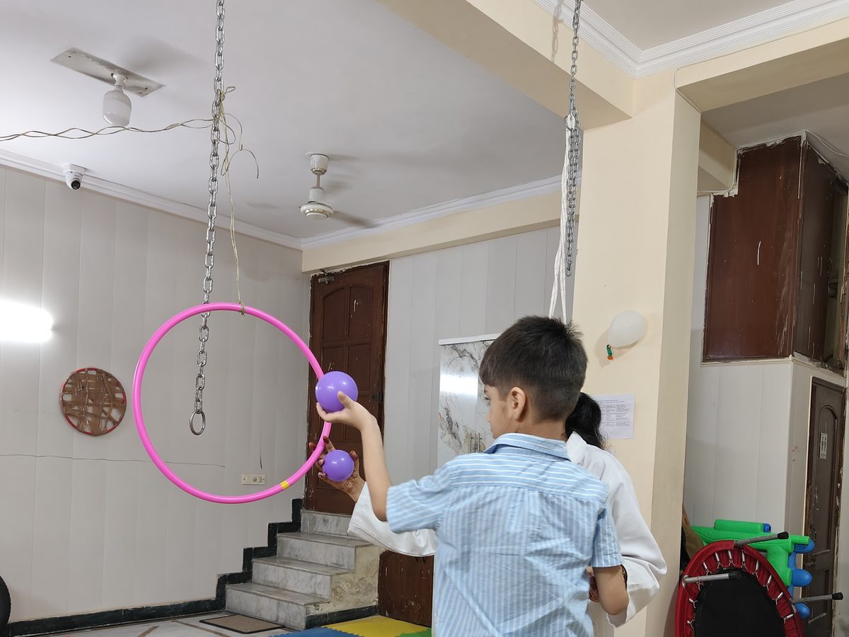 A child pushing soft balls through a hanging hoop, with a therapist close by, in the occupational therapy room at the Noida Centre.