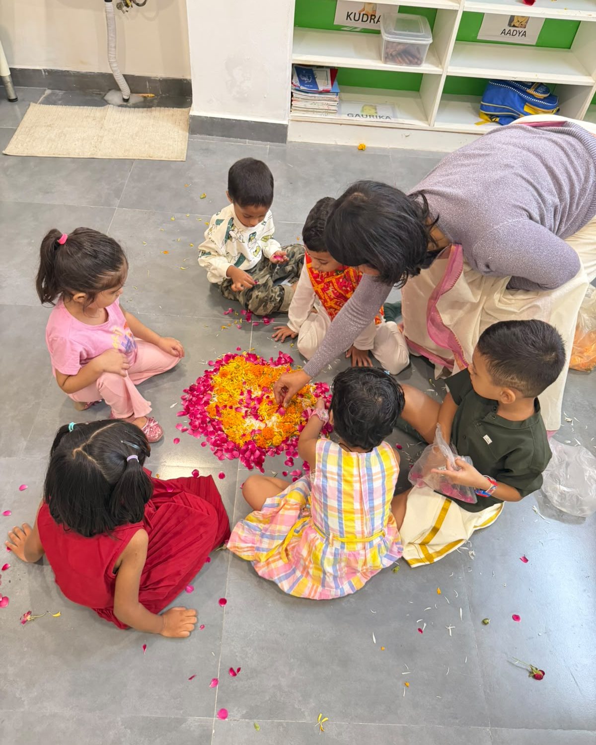 A teacher kneeling with a group of young children around a flower rangoli on the Early On classroom floor.