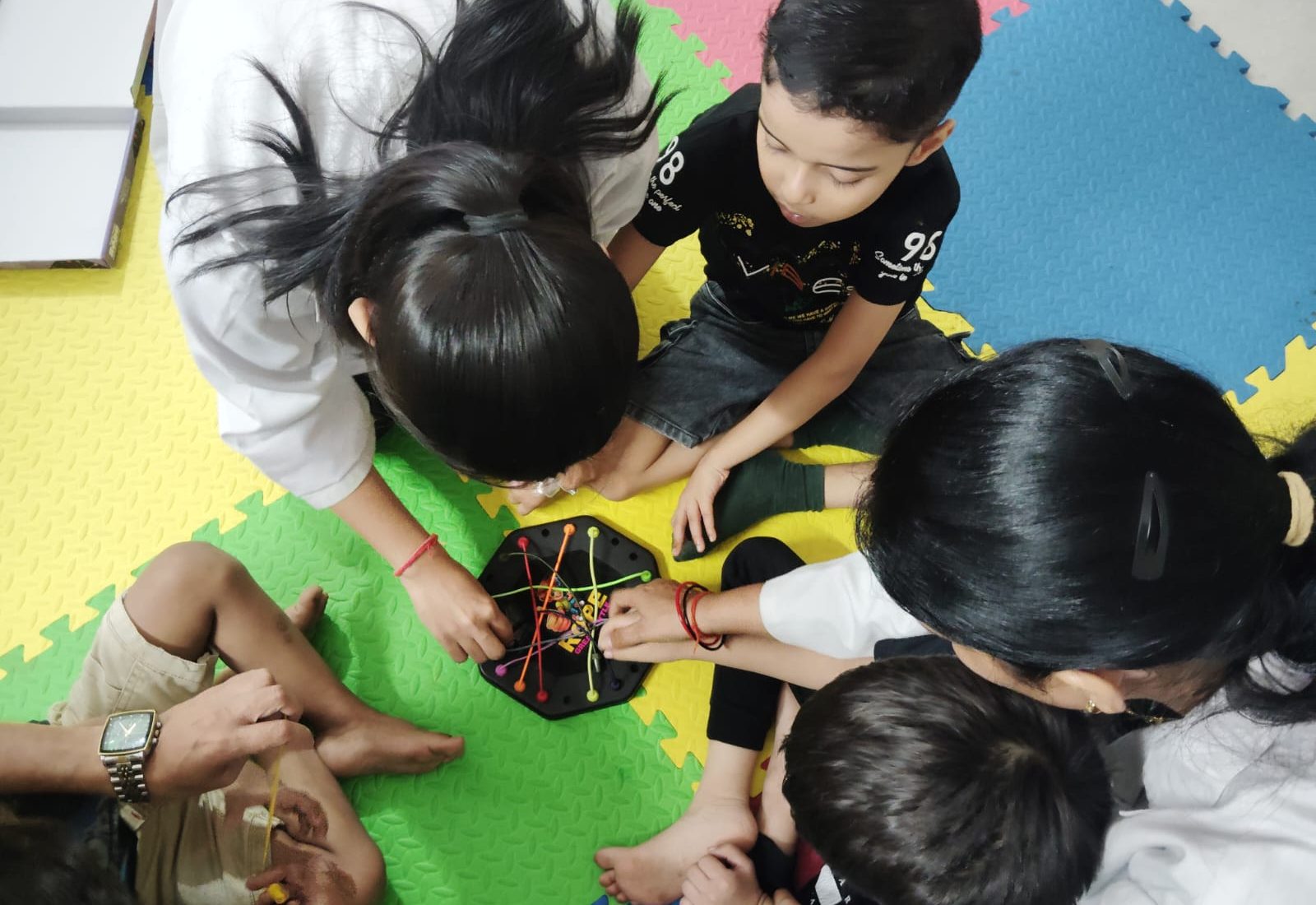Two CureAble therapists and a group of children gathered on a foam mat around a peg-board activity at the Noida Centre.
