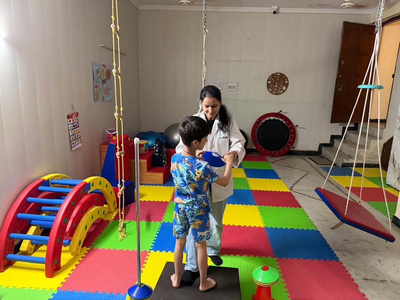 A CureAble therapist guides a child standing on a balance mat in a colourful occupational therapy room at the Noida Centre.