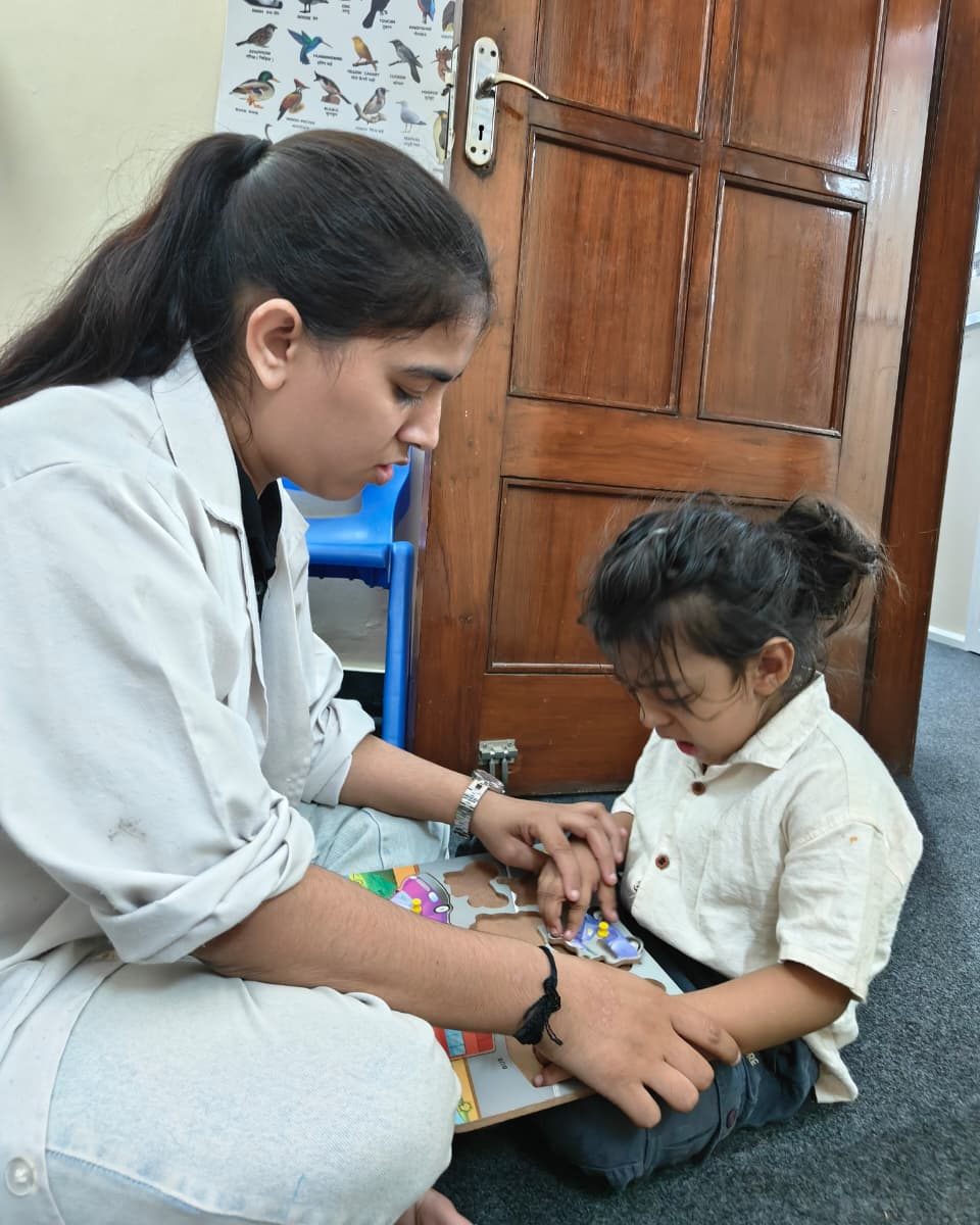 A CureAble therapist sits on the floor beside a young child, guiding the child's hands through a wooden puzzle.