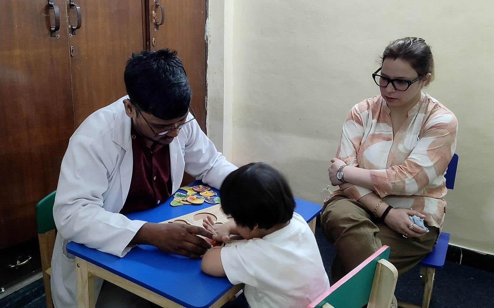 A CureAble clinician guides a young child through a shape puzzle while a parent looks on.