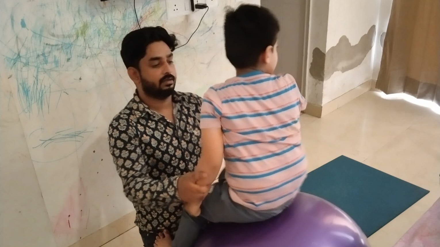 A therapist kneels beside a child sitting on a large therapy ball, holding the child's leg for balance, in a room at home.