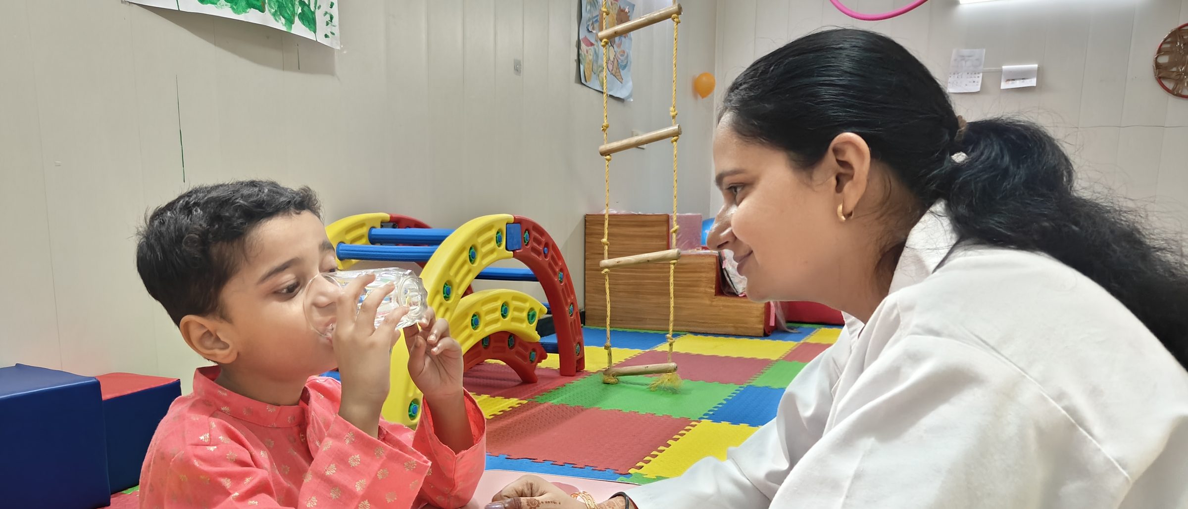 A smiling CureAble clinician sits across a small table from a young child drinking from a glass in a colourful therapy room.
