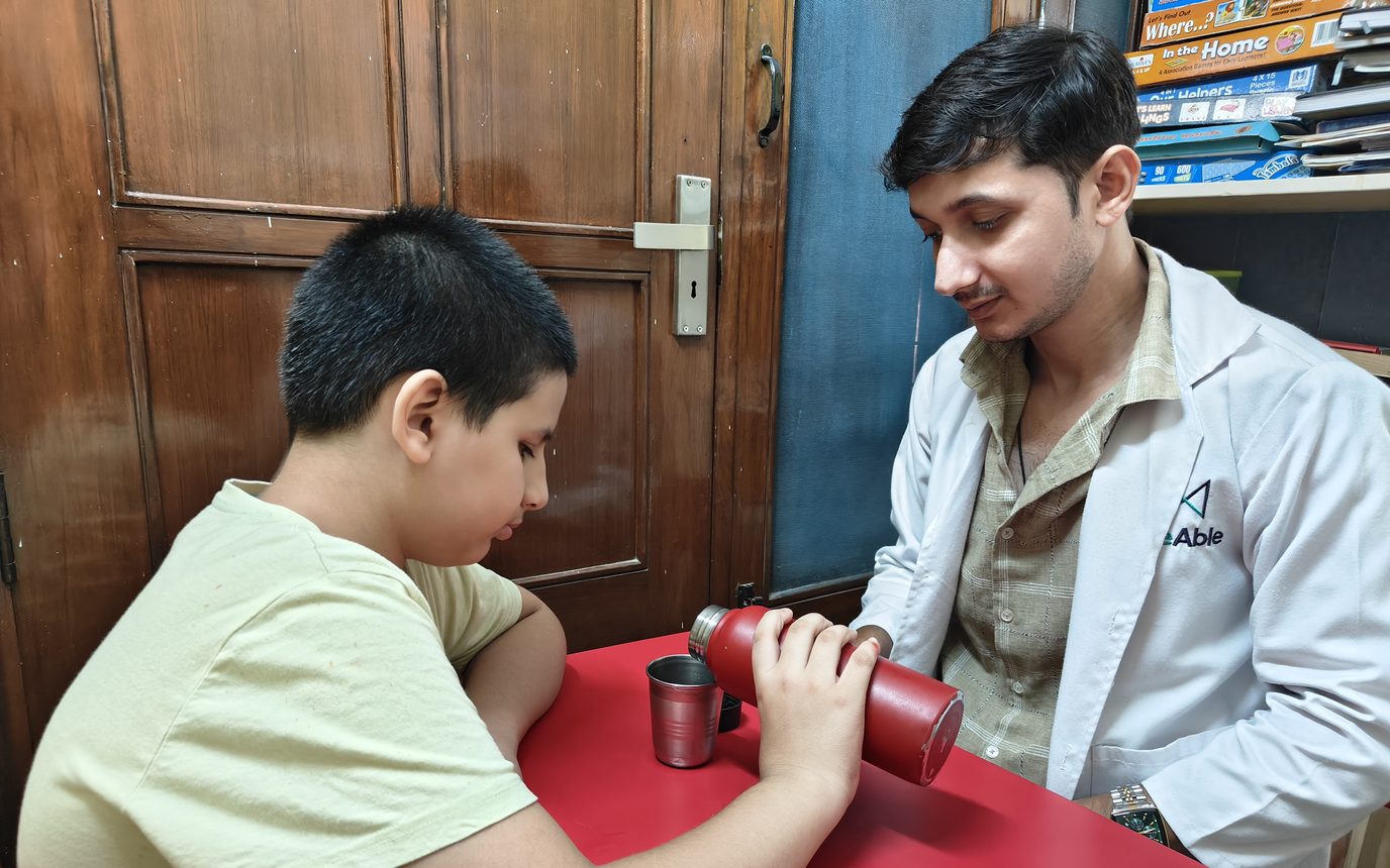 A CureAble therapist watches as a child pours from a flask into a cup at a red table at the Noida Centre.
