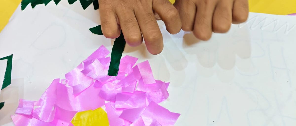 A child's hands pressing a paper stem onto a pink tissue-paper flower collage during an art and craft session at home.