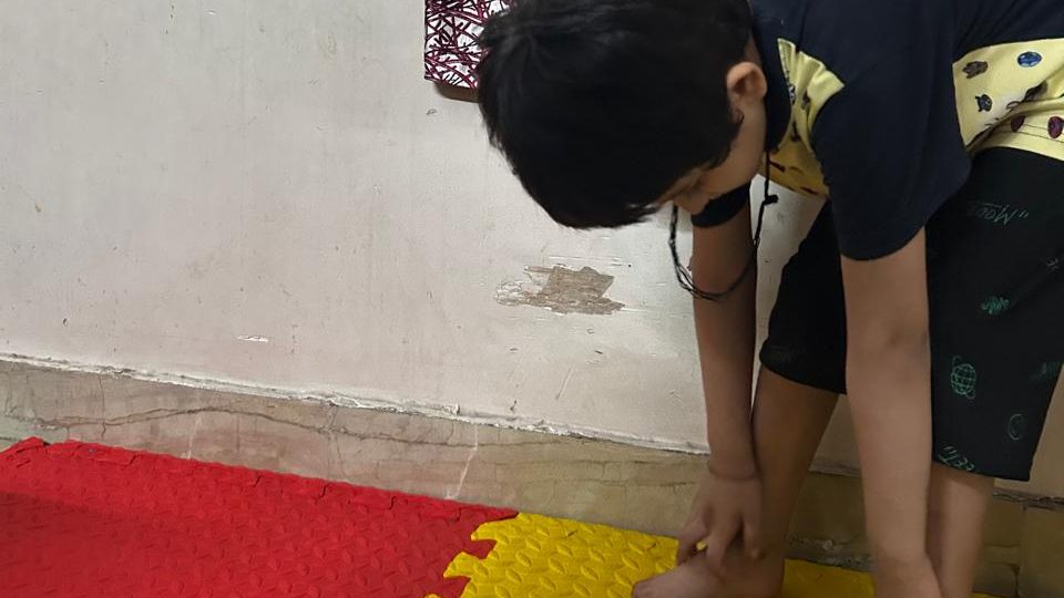 A child bends forward to reach their ankles on a foam play mat at home.