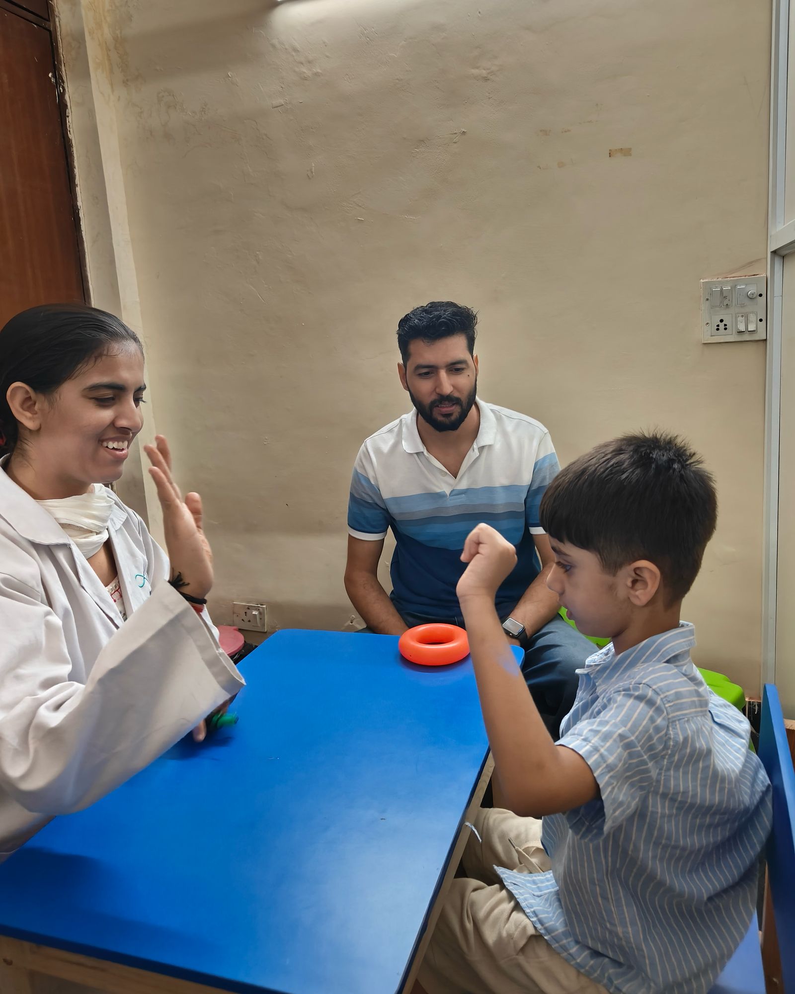 A smiling therapist, an adult and a child at a table during an activity in a CureAble therapy room.