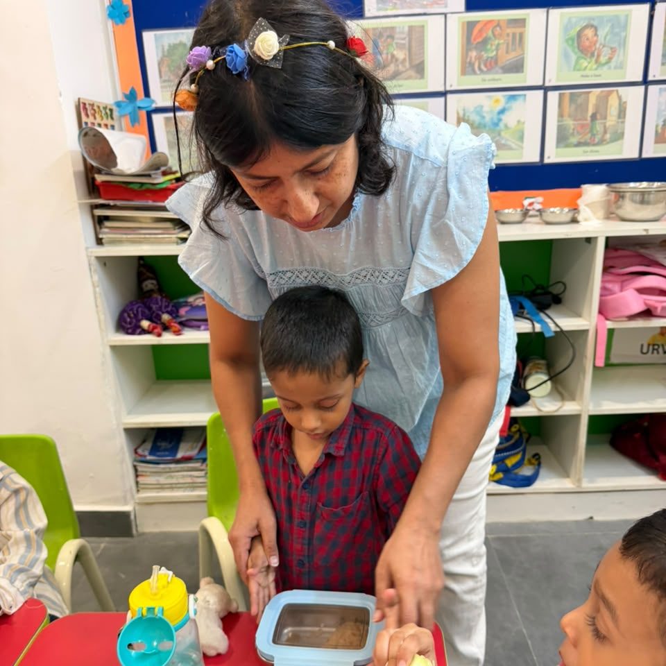 A teacher helping a young boy open his lunch box at the classroom table.