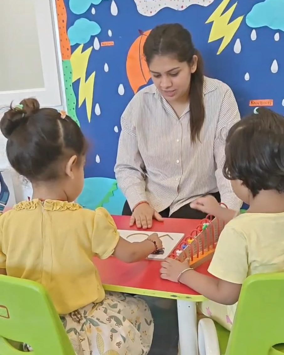 A teacher sitting at a classroom table with two young children working on a learning activity.