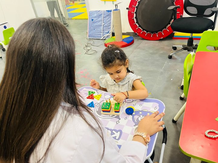 A therapist sitting with a young girl as she stacks shapes on a pegboard.