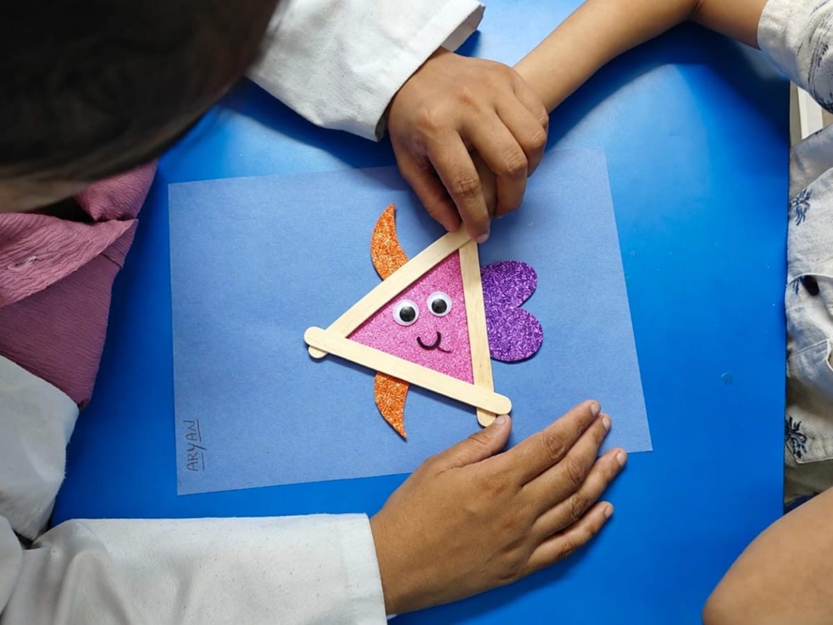A child's hands finishing a paper craft on a blue table, with a therapist alongside.