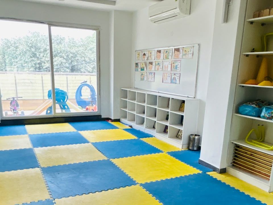 A bright EarlyOn room with blue and yellow floor mats, low shelves and a picture chart on the wall.