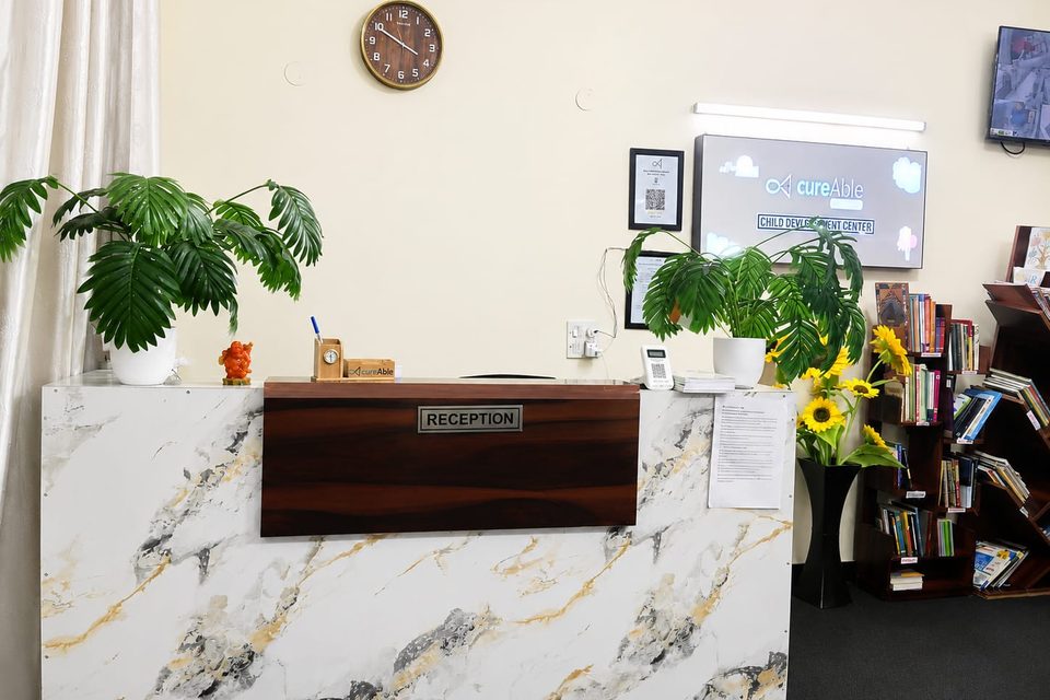The reception desk at CureAble Noida, with plants, a wall clock and a bookshelf.
