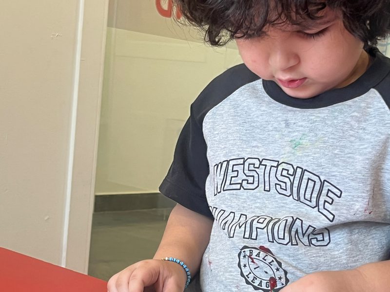 A young child concentrating on a painting activity at a classroom table.