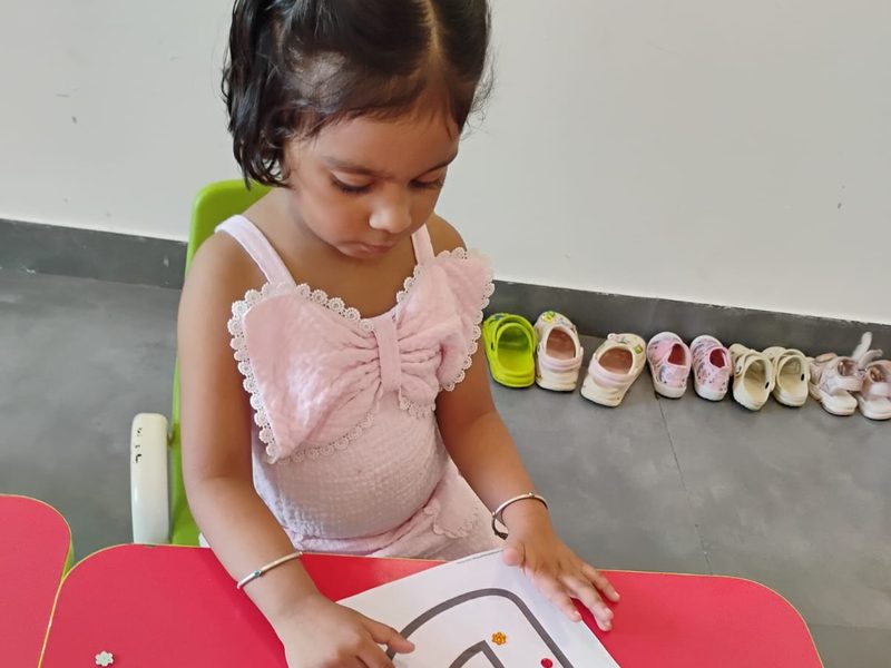 A girl placing coloured dots along a letter outline at a classroom table.