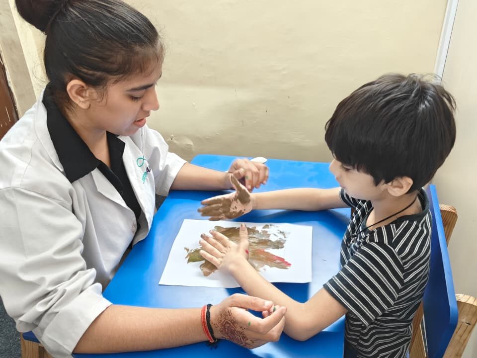 A therapist guiding a child's hands through a printing activity at a blue table at the Noida Centre.