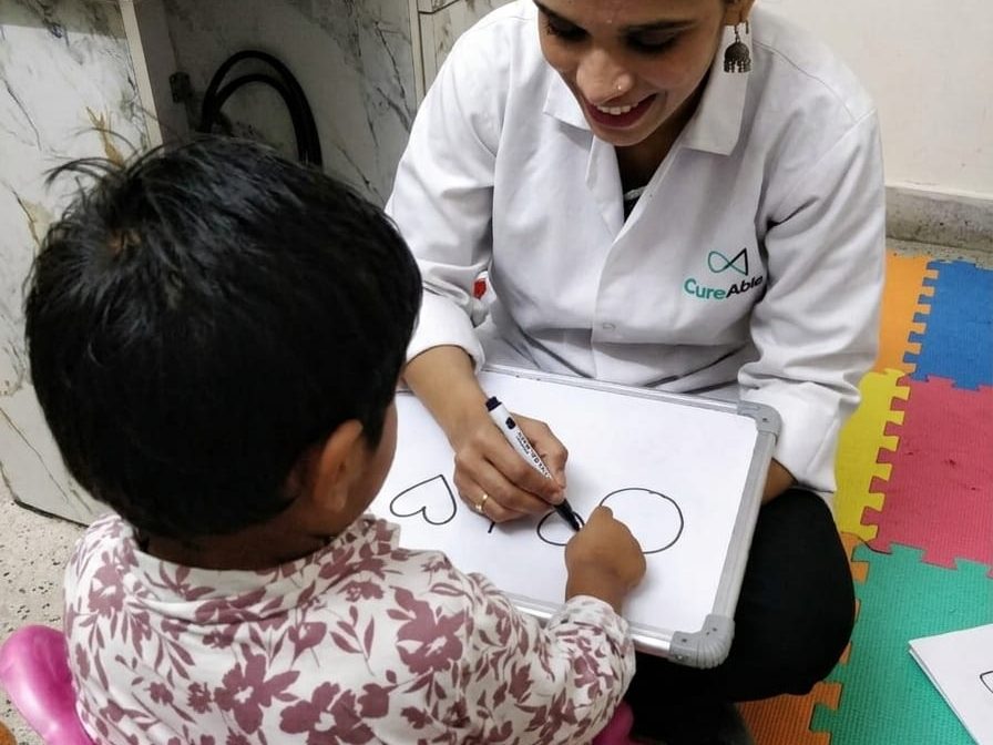 A special educator holding a small whiteboard while a child traces shapes on it at the Dwarka Centre.