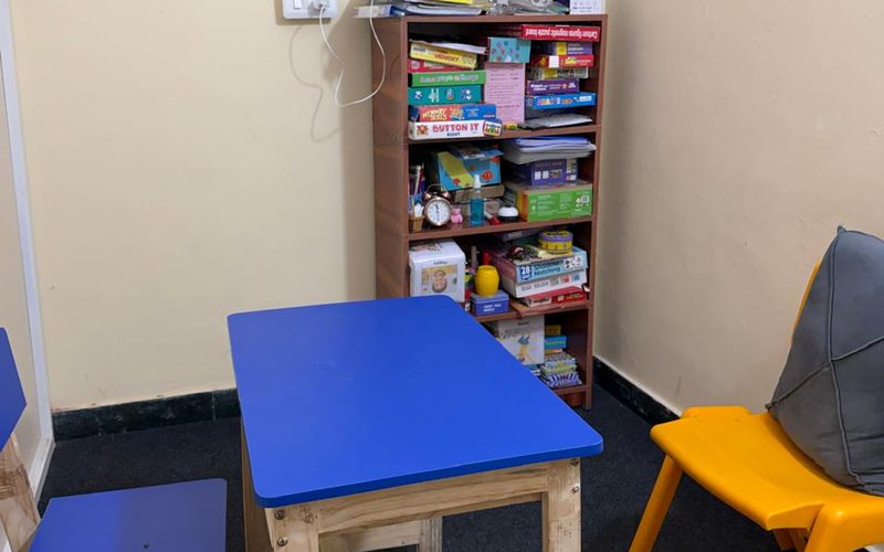 A small therapy room at CureAble Noida, with a blue table, a yellow chair and a shelf of games.